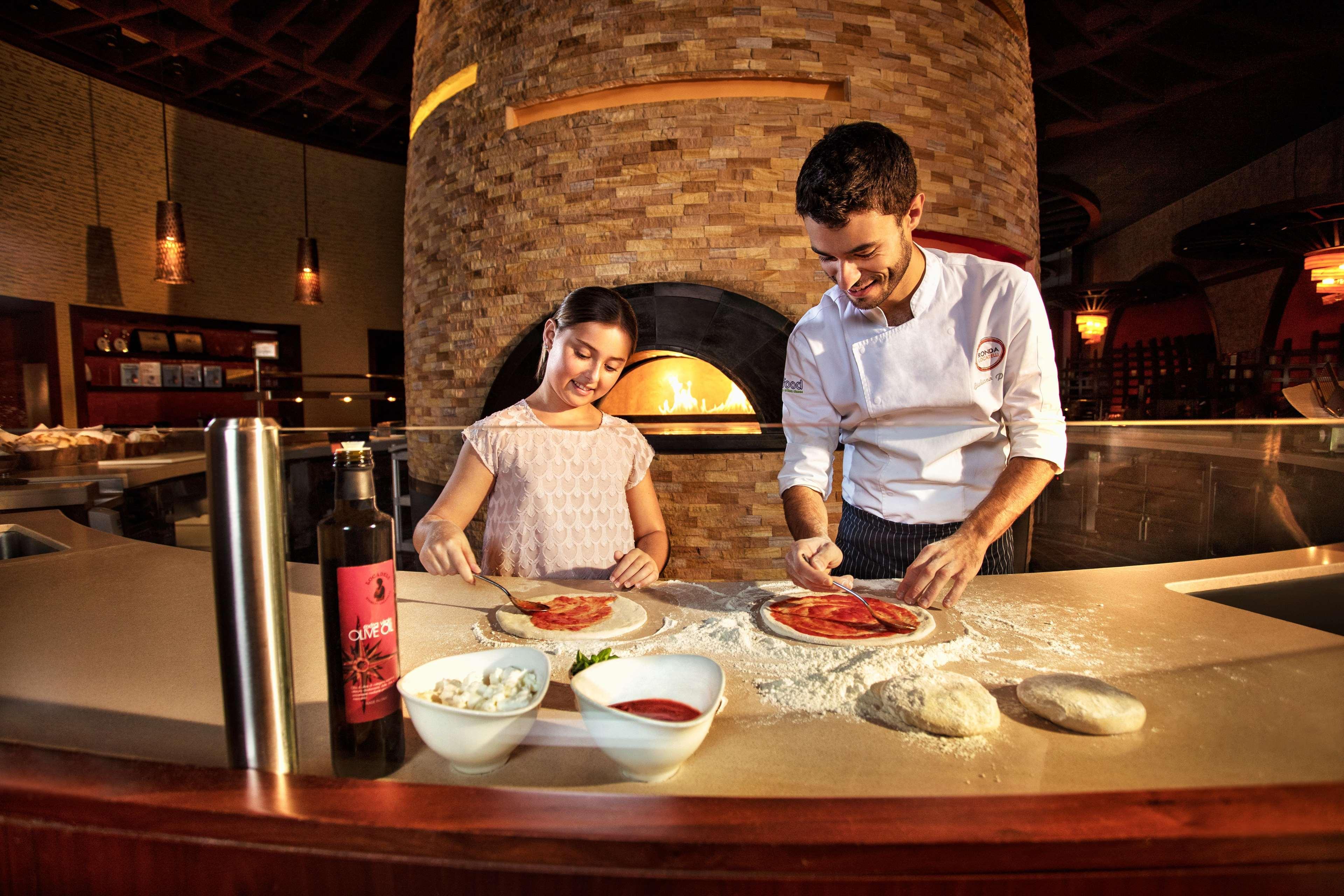 Atlantis, The Palm Dubai Exterior foto The photo shows a chef and a young girl working together in a kitchen setting. They are engaged in making pizza, with dough and various ingredients spread out on a counter. The girl is applying sauce or toppings to a pizza base while the chef, who ap